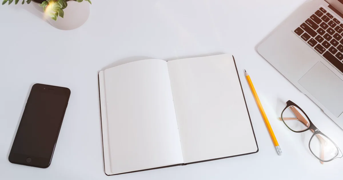 Světlý pracovní stůl s vypnutým mobilem, otevřeným čistým zápisníkem, tužkou, notebookem a brýlemi.
